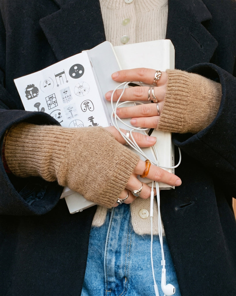 The Cashmere Fingerless Gloves in Raw, worn holding a notebook and earphones, with a navy coat