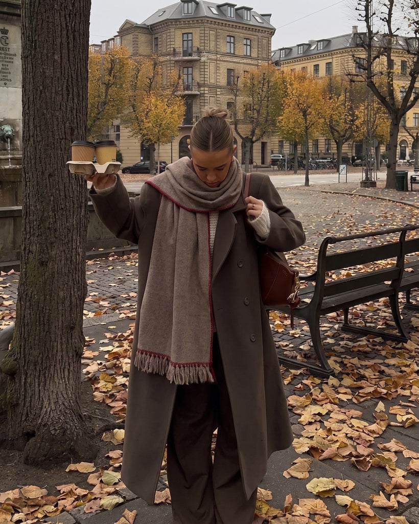 The Hohhot Heritage Wool Wrap in Raw, worn tucked under a brown coat in a park in autumn