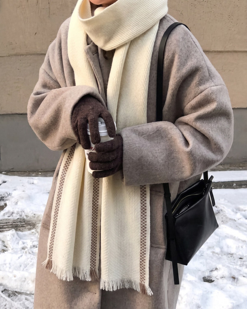 The Herringbone Wool Scarf in Ivory, worn hanging long over a taupe coat in the snow