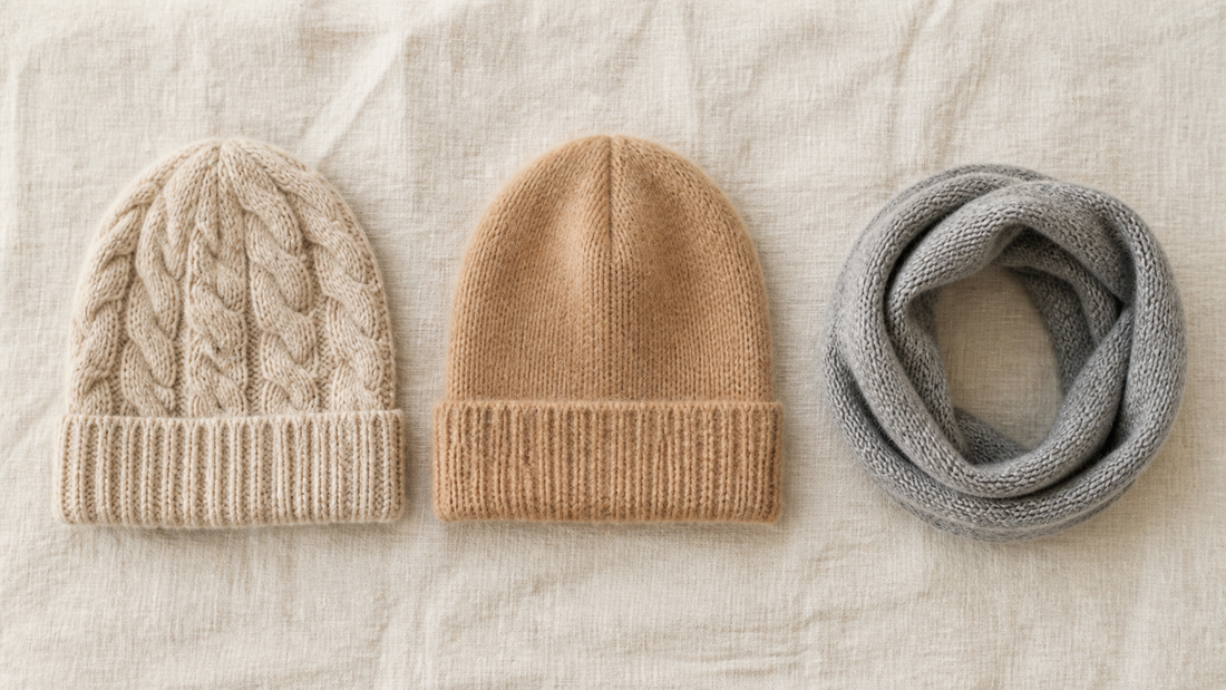 Flat lay of a cable-knit wool beanie, a smooth camel knitted beanie and a coiled grey knitted neck collar in a row on cream linen