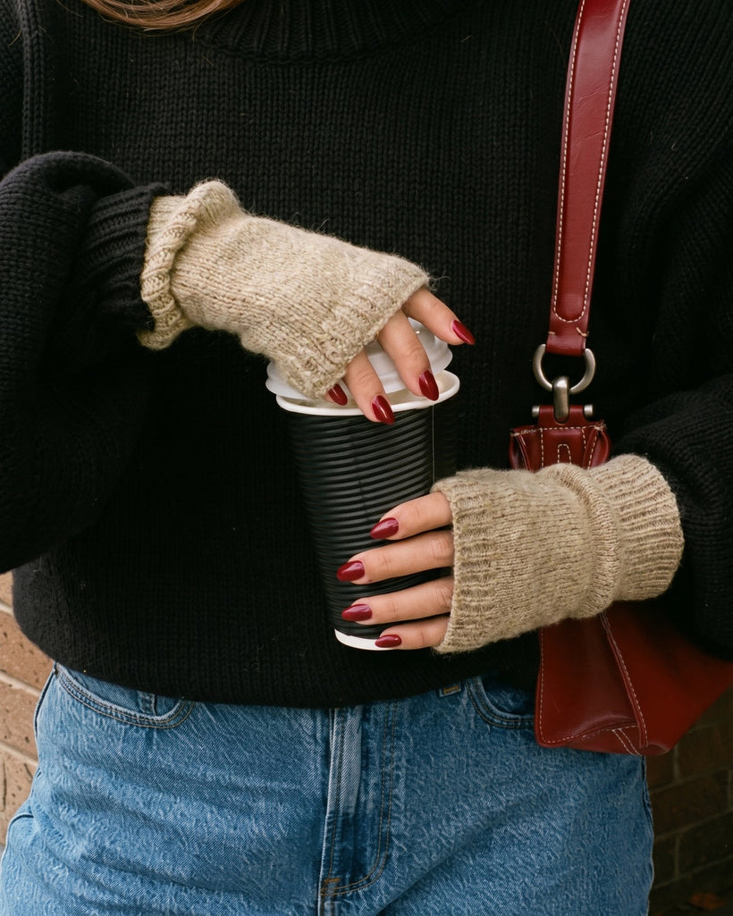 The Cashmere Fingerless Gloves in Raw, worn holding a coffee cup, with a black sweater and jeans