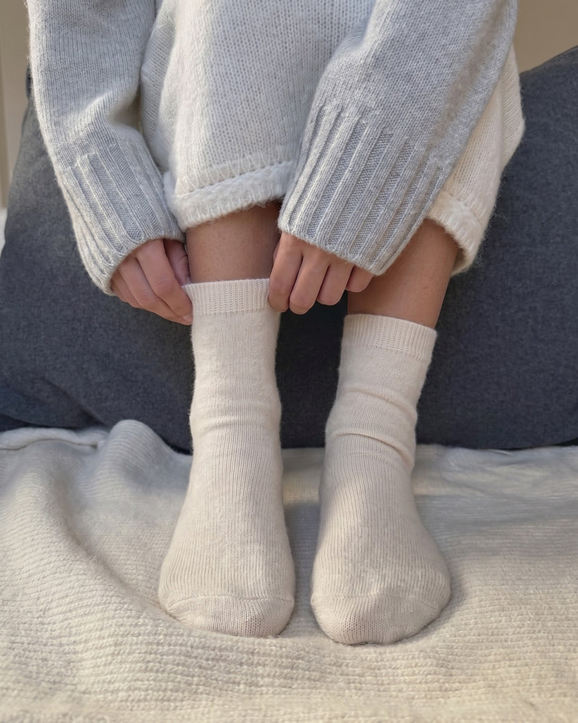 The 100% Cashmere Bed Socks in Beige, worn on a sofa with a grey knit, cuffs being pulled up