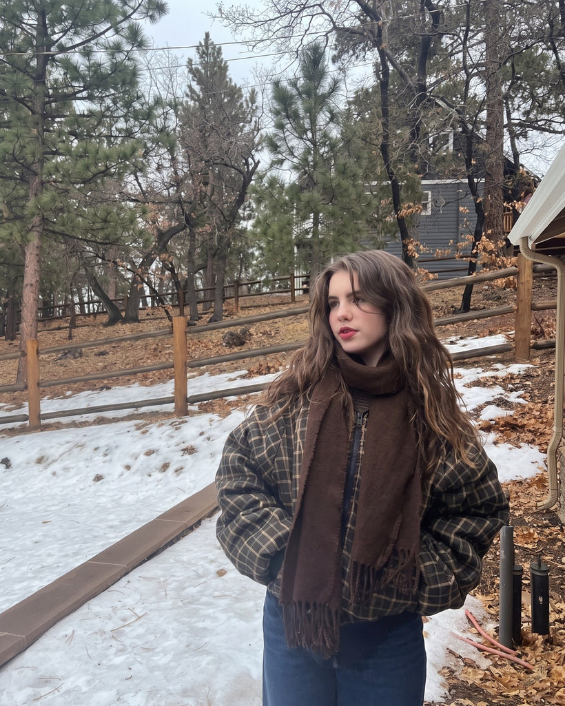 The Slim Wool Scarf in Espresso, worn knotted over a plaid jacket in the snow