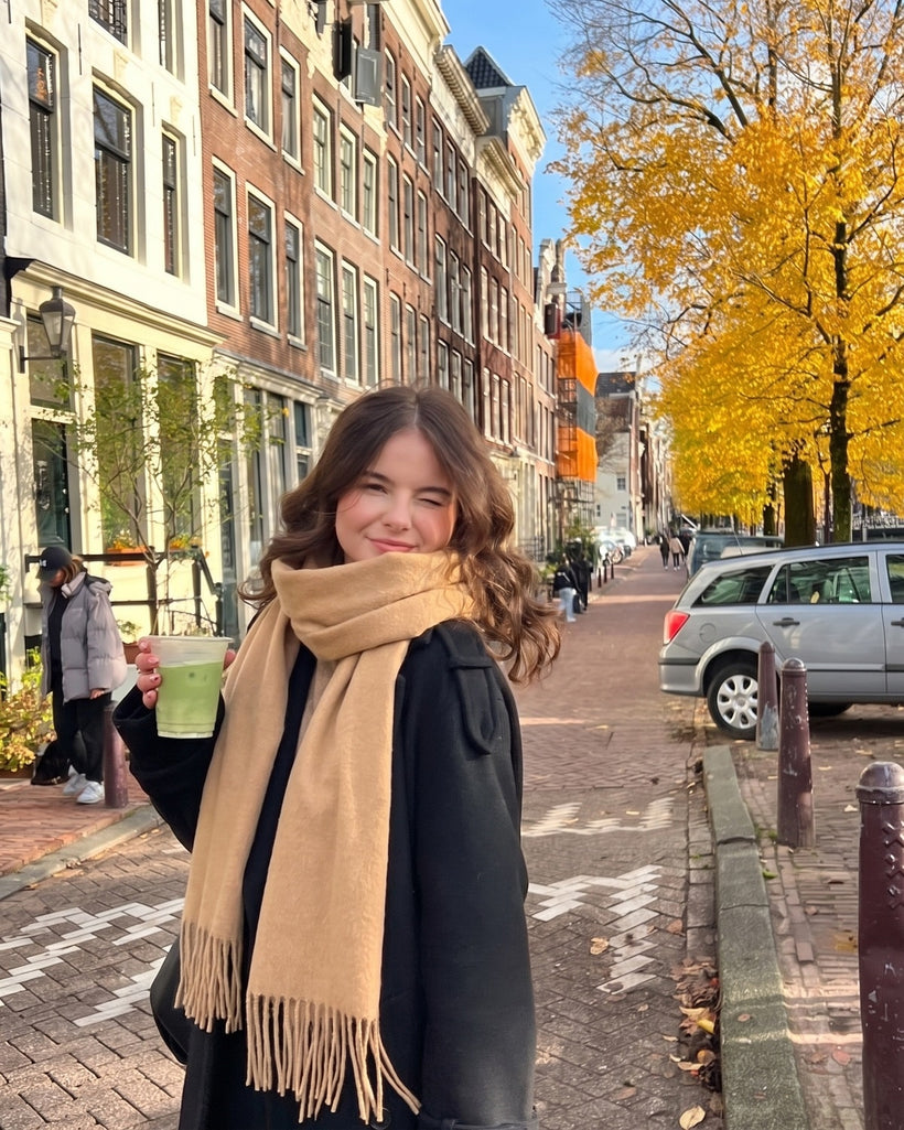 The Brushed Lambswool Long-Scarf in Camel, worn looped once over a black coat on a canal street