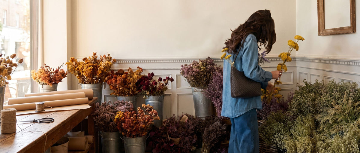 Woman in a denim shirt carrying the Wildfool woven leather shoulder bag with dried yarrow tucked inside, in a dried-flower shop